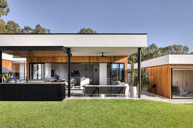 Exterior view of a contemporary single-storey residence with a broad flat roof and deep covered outdoor entertaining area. The facade combines white rendered surfaces, warm vertical timber cladding, slim dark metal posts and extensive glazing. A built-in black outdoor kitchen and barbecue occupy the covered patio, with outdoor seating facing a manicured lawn. A separate timber-clad pavilion or garage-like structure sits to the right.