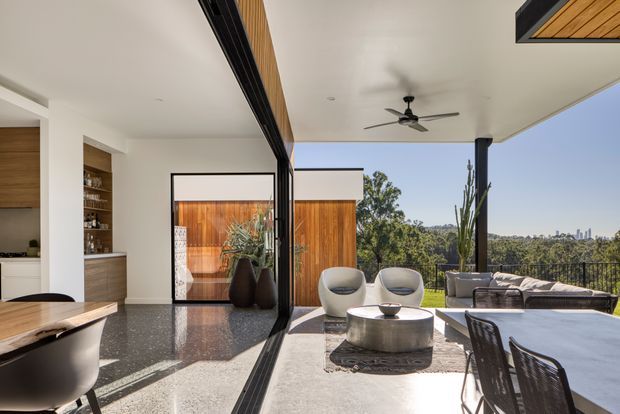A bright, contemporary open-plan interior extending to a covered outdoor terrace. The foreground shows a kitchen or dining area with a dark stone benchtop and timber cabinetry, while full-height black-framed sliding glazing opens to a lounge terrace with sculptural chairs, a low round coffee table, outdoor sofa, dining table, ceiling fan, and landscaped views. Warm timber screening and planter pots add contrast to the white ceiling and walls.