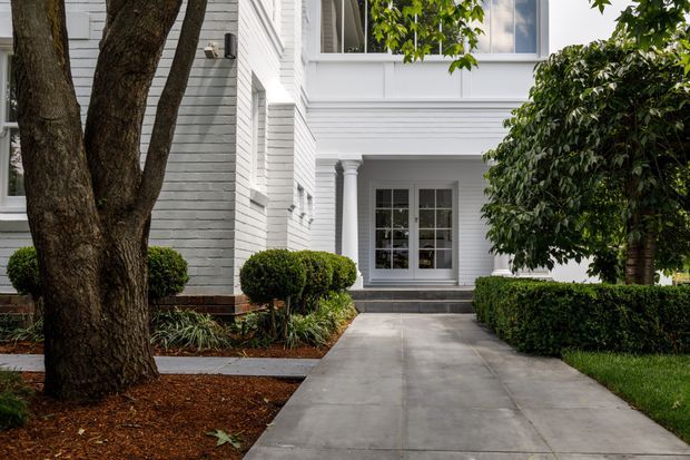 A formal residential exterior showing a white-painted brick and weatherboard-style facade with classical detailing, including fluted columns, decorative trim, multi-pane windows, and a covered entry. A straight concrete path leads through manicured hedges and clipped shrubs, with mature trees framing the approach.