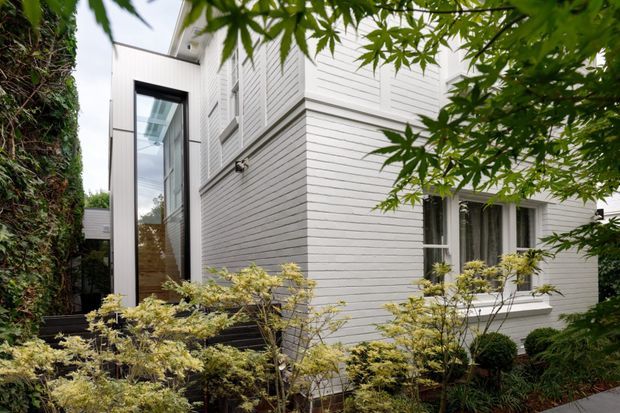 Exterior view of a contemporary multi-level residential building, partially screened by mature planting. The facade is clad in white horizontal weatherboards, with tall dark-framed glazing and white-framed windows. A narrow landscaped walkway and dense garden planting border the building.