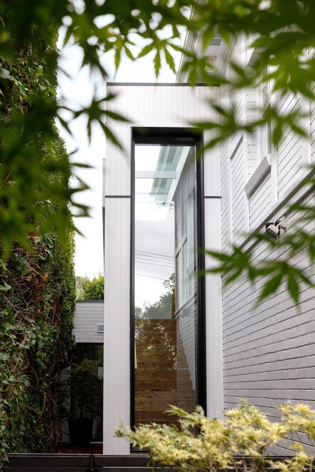 A close exterior view of a contemporary residential entry or glazed passage framed by dense greenery. The building features white horizontal brickwork, white vertical cladding, dark metal trim, and a very tall glazed doorway or window beneath a translucent roof canopy. The image is composed through foreground foliage, creating a layered garden setting.