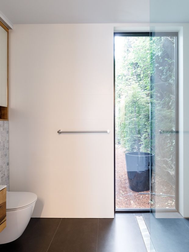 A compact, contemporary bathroom with a wall-mounted towel rail, white toilet, pale tiled wall, dark floor tiles, and a full-height glazed door or window opening onto a leafy outdoor area. Minimal built-in timber joinery is visible at the left edge, and the space has a clean, calm, light-filled design.