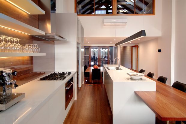 A sophisticated open-plan kitchen and dining area with white waterfall-edge island cabinetry, integrated gas cooktop, timber flooring, and a timber dining table with dark chairs. Warm illuminated display shelving with glassware lines the left wall, while a high ceiling reveals exposed dark timber roof structure through clerestory glazing. The space combines crisp white joinery with warm timber accents and strong linear lighting.