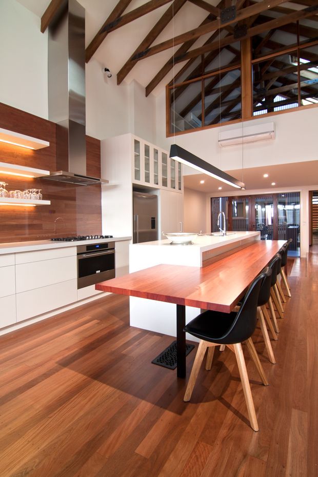 A spacious open-plan kitchen and dining area with a double-height ceiling. The kitchen features white flat-panel cabinetry, a large white island with integrated sink, timber benchtops and wall cladding, stainless steel appliances, and a long timber dining table with black bar stools. Exposed dark timber roof trusses and a glazed upper-level void add strong architectural character. Warm timber flooring runs throughout, with contemporary pendant and recessed lighting.