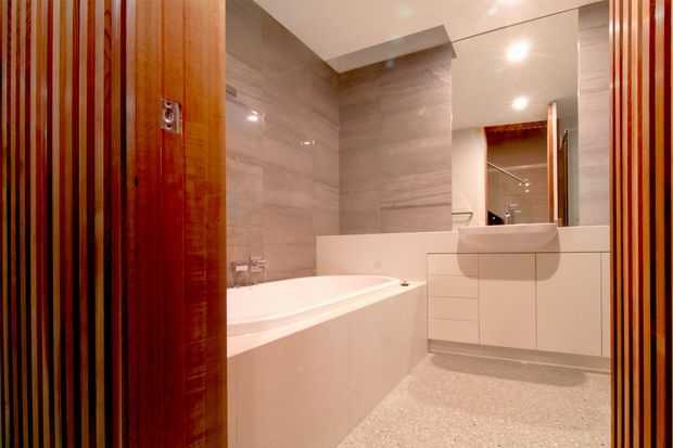 Contemporary bathroom viewed through a timber slatted doorway, featuring a built-in white bathtub, wall-mounted white vanity with integrated basin, large mirrored cabinet, recessed ceiling lighting, and grey stone-look wall tiles with a terrazzo-like floor.