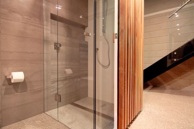 Modern bathroom with a frameless glass walk-in shower, large-format taupe wall tiles, a linear floor drain, and a textured light stone or terrazzo-style floor. A vertical timber slat partition partially screens the shower from an adjacent area, with a black stair or balustrade visible beyond.