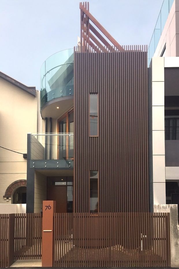 A narrow contemporary multi-storey urban residence viewed from the street, featuring a tall dark timber-look vertical slatted facade and matching privacy gate. Curved glass balcony balustrades and recessed glazing sit alongside a pale rendered neighbouring structure, while a projecting upper timber screen adds a strong vertical architectural feature. The facade combines warm brown slats, light wall panels, and transparent glass in a compact infill-style composition.
