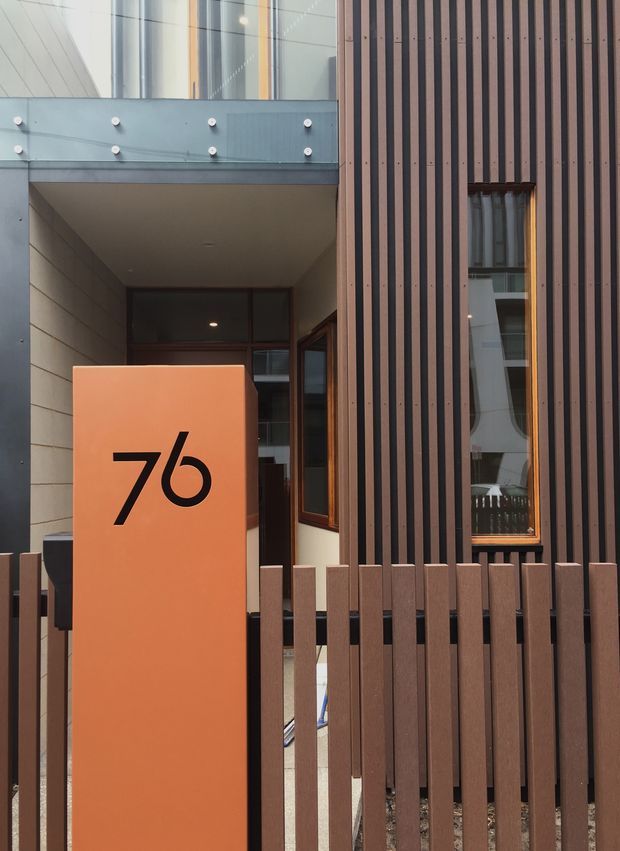 Close-up exterior view of a contemporary building entrance. The facade combines vertical dark timber-look battens, large glazed windows with dark frames, and pale grey wall panels. A glass-balustraded upper-level walkway projects over the recessed entry, while an orange address pillar marked “76” stands behind a matching vertical-batten fence.