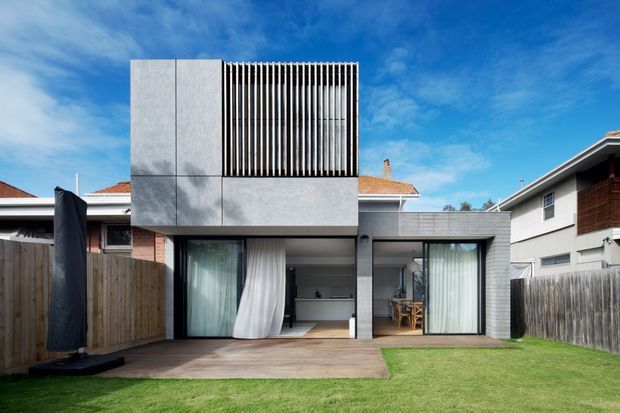 Rear exterior view of a contemporary two-storey residential extension with a strong geometric form. The upper level is clad in smooth light-grey panels and screened with closely spaced vertical slats, while the ground floor has expansive glazed openings with curtains leading to a timber deck and lawn. Adjacent suburban homes and boundary fencing are visible.