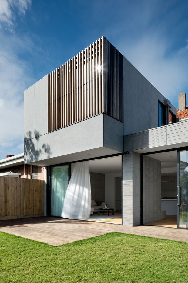 Exterior view of a contemporary multi-level residential extension or townhouse, with a clean rendered upper volume, prominent vertical timber screening, extensive ground-floor glazing, a pale tiled facade, and a timber deck opening onto a lawn. The building combines rectilinear forms with a visible adjacent pitched terracotta-tiled roof and neighbouring residential context.