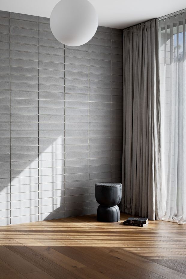 A minimalist interior corner with a grey concrete-block feature wall, warm timber flooring, full-height layered curtains, and a large spherical pendant light. A small sculptural black side table and books sit near the window, with strong natural sunlight casting shadows across the wall and floor.