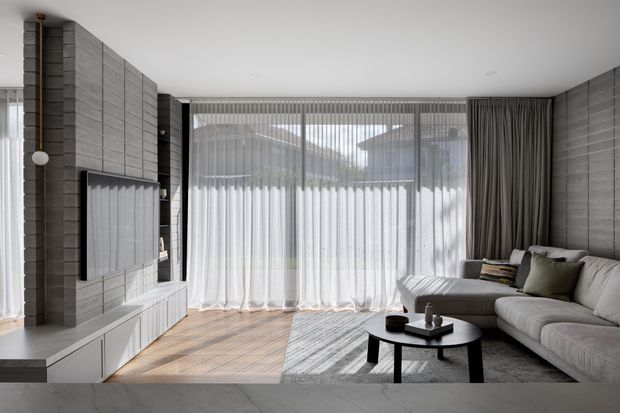 Contemporary living room with a large grey tiled feature wall incorporating a wall-mounted television and low white built-in cabinetry. Floor-to-ceiling glazed doors with sheer curtains bring in strong natural light, while a grey sectional sofa, round black coffee table, and layered neutral furnishings create a calm, refined interior. Light timber flooring and vertical grey wall cladding provide textural contrast.