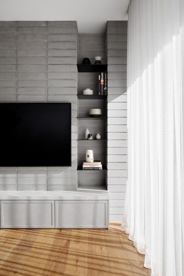 A bright, minimalist living room media wall with exposed grey concrete blockwork, a wall-mounted black television, and a narrow black open shelving niche displaying books and small decorative objects. White built-in cabinetry runs along the base, while sheer full-height curtains filter strong sunlight from the right. Warm timber flooring and striped shadows add contrast to the otherwise monochromatic interior.