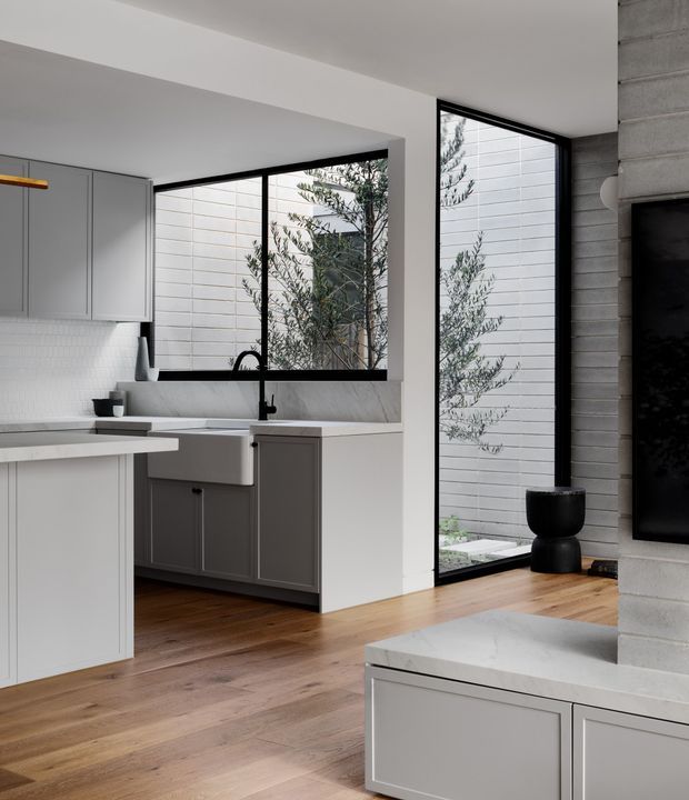 A refined contemporary open-plan kitchen viewed toward a living area, with pale grey flat-panel cabinetry, a white tiled splashback, a white farmhouse sink, dark fixtures, and a light stone benchtop. Warm timber flooring contrasts with black-framed floor-to-ceiling glazing and a textured grey masonry feature wall. A narrow planted courtyard is visible beyond the windows, while a television and built-in cabinetry appear at right.