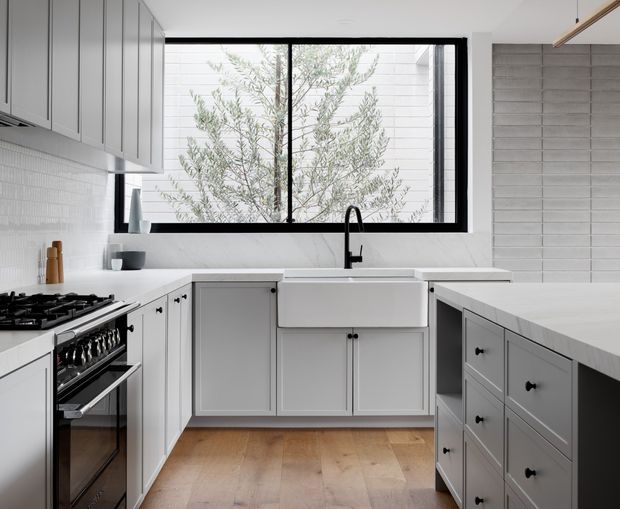 Bright, compact contemporary kitchen with a U-shaped layout and central island. The space features light grey Shaker-style cabinetry with black knobs, white benchtops, a large white farmhouse sink, black tapware, a freestanding black range cooker, pale timber flooring, and a large black-framed window overlooking a planted courtyard. A horizontal grey tiled wall provides a subtle feature backdrop.