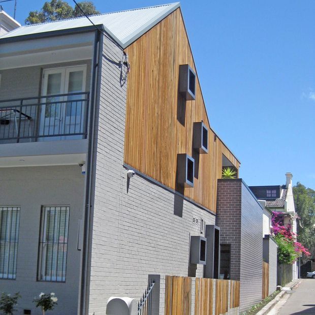 Street-level exterior view of a narrow contemporary multi-storey residential building in an urban setting. The facade combines vertical warm timber cladding, pale painted brickwork, dark metal window surrounds, projecting box windows, a corrugated metal gabled roof, and a small balcony with dark metal railing. Adjacent traditional residences and landscaped planting are visible along the street.