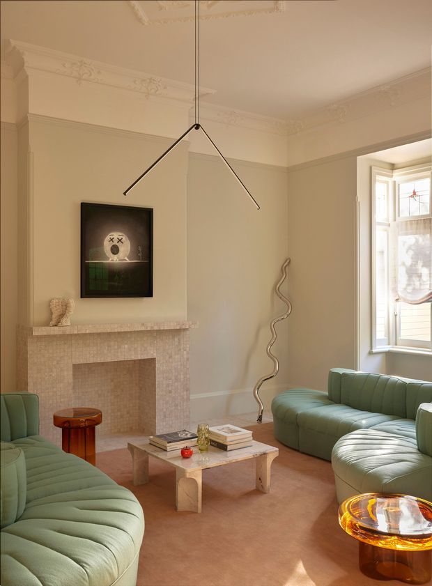 Elegant living room in a period home with ornate plaster ceiling mouldings, a tiled fireplace surround, tall bay window, and sculptural contemporary pendant light. The room is furnished with curved sage-green channel-tufted sofas, a marble coffee table, a small timber side table, and contemporary artwork. The palette is warm and softly muted, combining heritage detailing with modern furnishings.