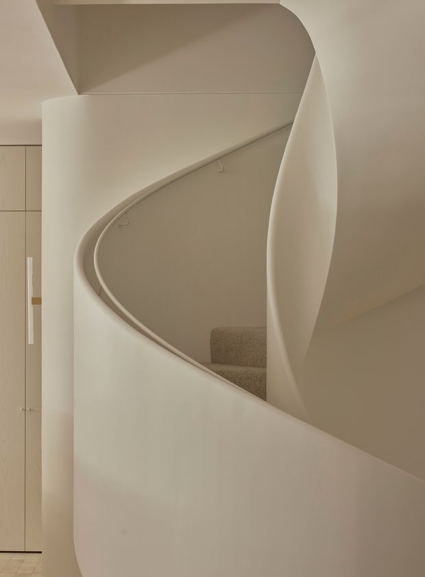 A softly lit interior view of a sculptural curved staircase. Smooth off-white plaster walls and a sweeping balustrade create an organic, minimalist form, with a small section of neutral carpeted treads visible behind the curve and pale built-in cabinetry at left.