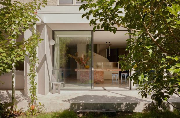 A leafy residential courtyard view looking through full-height glazing into a contemporary open-plan interior. The pale rendered facade and terrace frame a compact kitchen or bar area with a patterned stone island, timber cabinetry, pendant lights, and stools. Mature planting partially screens the building, creating dappled sunlight and a calm indoor-outdoor connection.
