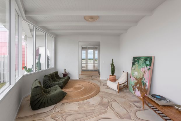 Bright, airy modern living room with full-height glazing along one side, white painted ceiling beams and walls, sculptural dark green lounge seating, a large round natural-fibre rug, a timber-framed lounge chair, indoor plants, artwork, and a low timber console. The room opens toward a further glazed area with an outdoor view, and features distinctive light stone-patterned flooring.