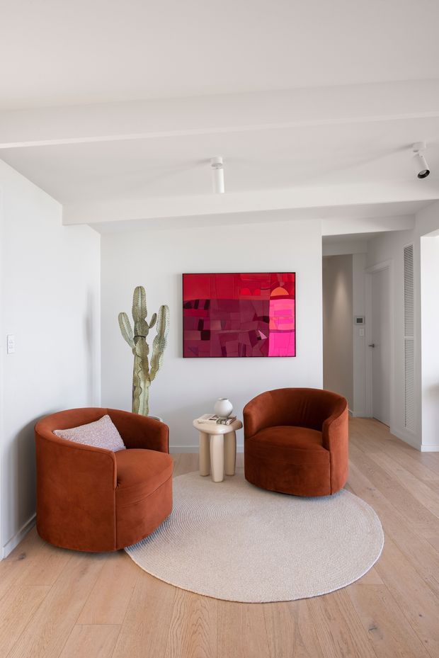 A bright, compact sitting area with two rust-orange rounded lounge chairs arranged around a small light-colored side table. A pale oval rug sits over light timber flooring, while a vivid abstract red and pink artwork forms the main wall feature. A tall cactus and minimal decorative objects add organic detail. White walls, a simple flat ceiling with recessed and track lighting, and a hallway or entry opening create a clean contemporary interior.