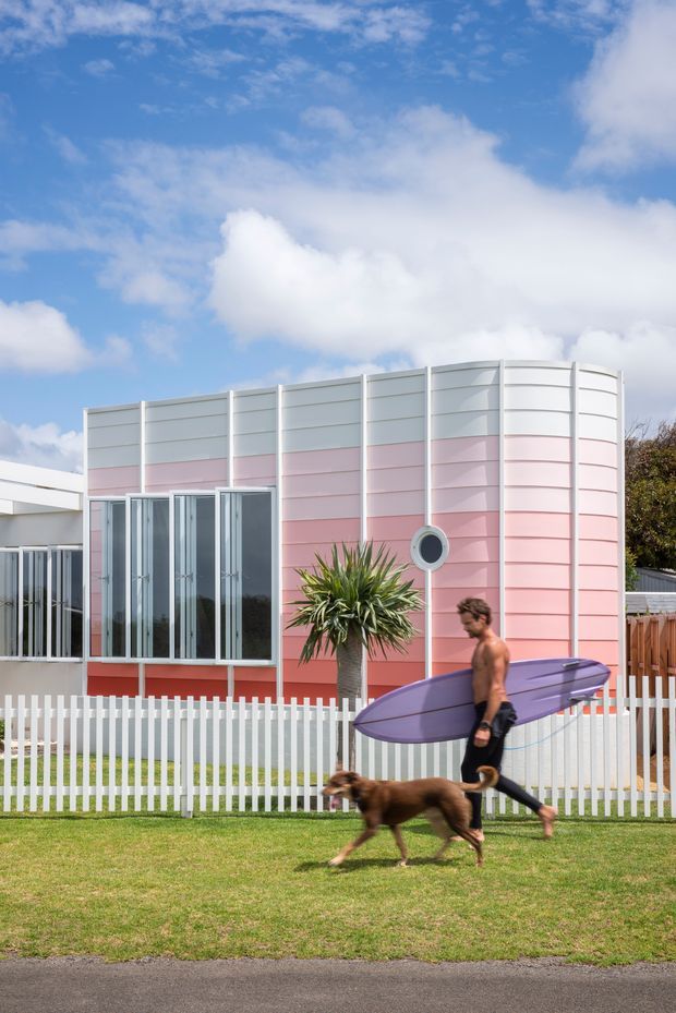Exterior view of a distinctive small residential building with a curved end wall clad in horizontal pastel pink and white panels. Tall glazed doors and windows line the facade, with a circular porthole-style window and a white picket fence in front. A grassy verge and paved street occupy the foreground, while a person carrying a surfboard and walking a dog passes by.