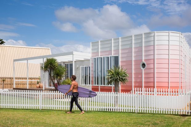 Exterior view of a distinctive low-rise coastal-style building with a rounded, horizontally panelled pink-and-white facade, extensive vertical glazing, and a white picket fence. A separate light-coloured gabled volume and simple covered outdoor structure sit to the left, with spiky ornamental plants in the foreground. A person carrying a surfboard reinforces the beachside character, though the building and landscape are the primary subjects.