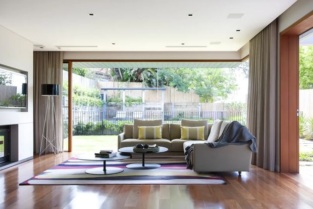 Bright contemporary living room with expansive floor-to-ceiling glazing opening to a landscaped backyard. The room features polished timber flooring, a white flush ceiling with recessed downlights, full-height taupe curtains, warm timber-framed glazing, a large neutral sectional sofa, sculptural low coffee tables, and a colourful geometric area rug. A television and media joinery are visible at left, while the garden and outdoor setting are seen through the glazed wall.