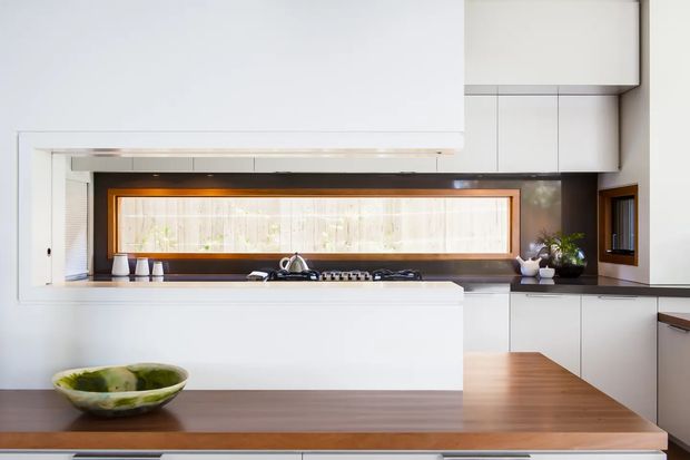Bright contemporary kitchen with extensive white cabinetry, a large central island, and a warm timber benchtop. A long horizontal window with a dark timber frame forms a feature backsplash above the cooktop, while potted plants and a decorative bowl add subtle natural accents.