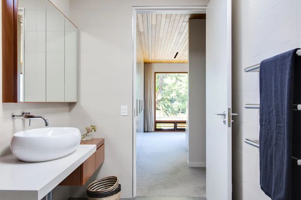 Bright contemporary bathroom with a white vessel basin on a white vanity, wall-mounted mirrored cabinetry, chrome tapware, pale tiled walls, and a dark towel on a heated towel rail. An open doorway leads to a carpeted room with timber-lined ceiling and a large window overlooking greenery.