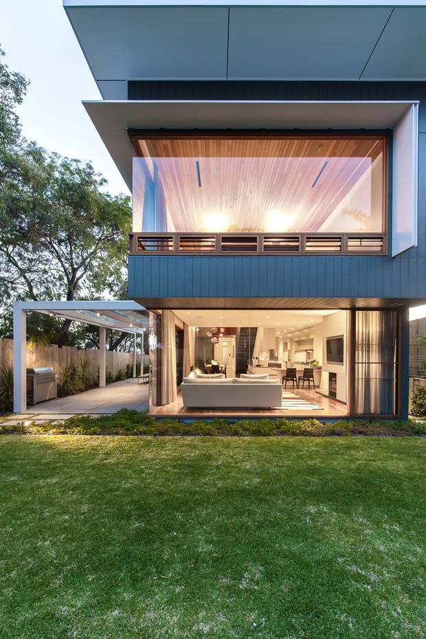 A modern two-storey residential house viewed from the landscaped backyard. The home features strong horizontal roof overhangs, dark blue-grey vertical cladding, extensive floor-to-ceiling glazing, and a projecting upper-level sunroom or balcony with timber-lined soffit and horizontal balustrade. The lower level opens broadly to an airy living and kitchen area, while a covered outdoor entertaining zone with barbecue sits to the left. The architecture has clean geometric lines, warm timber detailing, and a close relationship between indoor and outdoor spaces.
