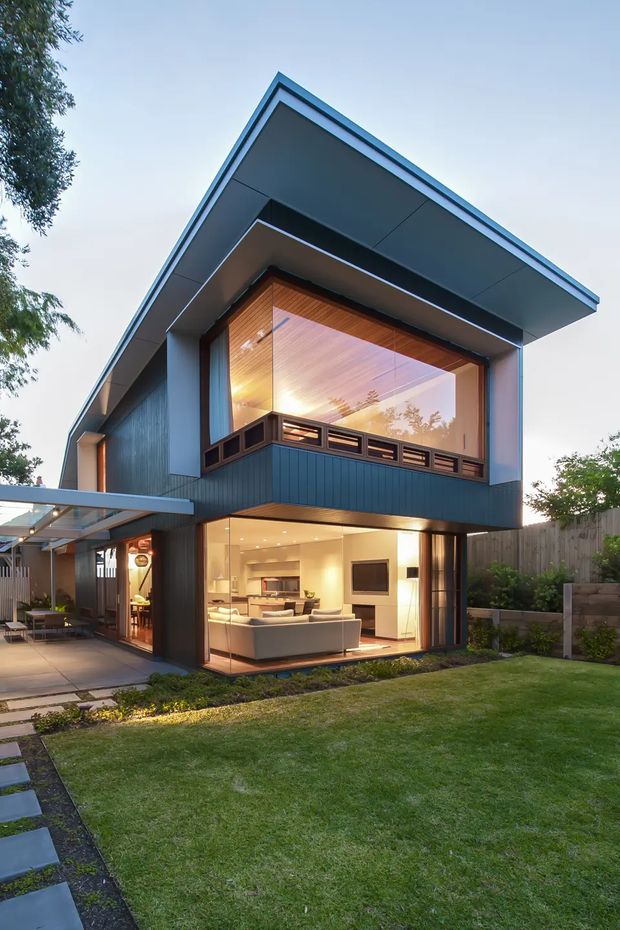 Exterior view of a contemporary two-storey house at dusk, featuring dark vertical cladding, a prominent projecting gabled roof, expansive glazed openings, a timber-lined upper-level balcony, and an illuminated open-plan living area. A covered outdoor area, paved walkway, lawn, planting, and boundary fencing surround the home.