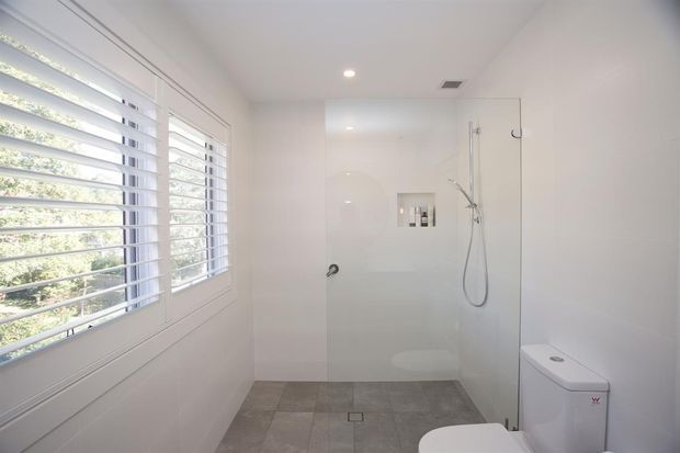 Bright, minimalist bathroom with white tiled or panelled walls, a frameless glass walk-in shower, recessed shower niche, wall-mounted shower fittings, and a white toilet. A large shuttered window provides natural light, while grey floor tiles add subtle contrast.