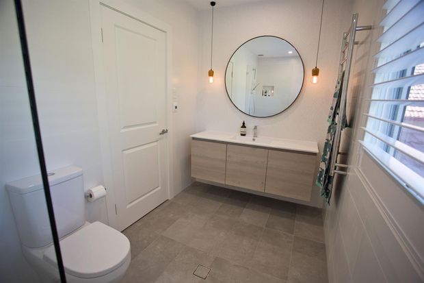Bright contemporary bathroom with a wall-hung timber-look vanity, white benchtop, round black-framed mirror, pendant lights, grey tiled floor, white toilet, and a glass shower enclosure. White plantation shutters provide filtered daylight.