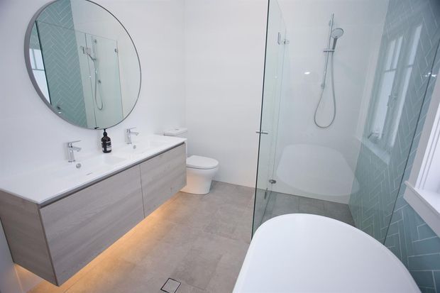 Bright contemporary bathroom featuring a floating double vanity with pale wood-grain cabinetry and integrated under-cabinet lighting, a large round mirror, wall-hung toilet, frameless glass shower enclosure, freestanding white bathtub, and pale blue-green geometric wall tiles. The room has a clean, airy finish with light neutral flooring and white sanitaryware.