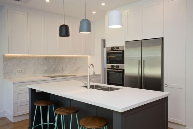 Bright, contemporary kitchen featuring a large white island with dark cabinetry, integrated sink and tap, white shaker-style cabinetry, marble-look splashback, stainless-steel double-door refrigerator, stacked ovens, and three pendant lights. Timber-topped stools with coloured metal legs provide contrast beneath the island.