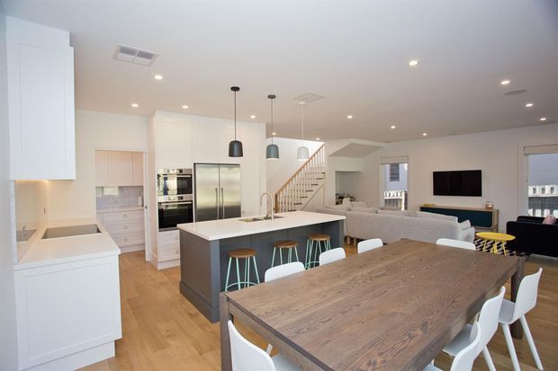 Bright open-plan kitchen, dining and living area with white cabinetry, stainless-steel appliances, a central island with pendant lights and stools, timber flooring, a large rustic timber dining table, and a staircase leading to an upper level.