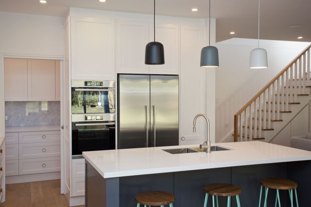 Bright contemporary kitchen with white full-height cabinetry, integrated stainless-steel ovens and double-door refrigerator, a large white island with undermount sink and curved faucet, and dark blue island cabinetry. Three pendant lights hang above the island, while a timber staircase and a walk-in pantry or utility area are visible beyond.
