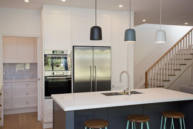 A bright contemporary kitchen with a large white island, dark blue base cabinetry, integrated stainless-steel appliances, and three contrasting pendant lights. A pale built-in pantry and countertop are visible to the left, while a timber-railed staircase rises behind the kitchen.