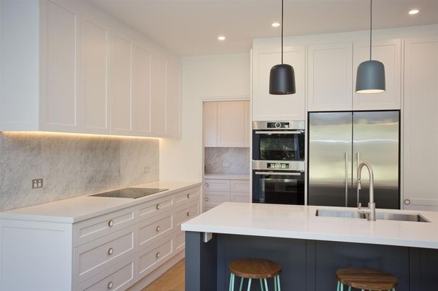 Bright, refined contemporary kitchen with extensive white panelled cabinetry, integrated double ovens, stainless-steel French-door refrigerator, pale stone benchtops and splashback, and a dark island with seating. Pendant lights and under-cabinet lighting provide warm contrast, while timber flooring adds natural warmth.