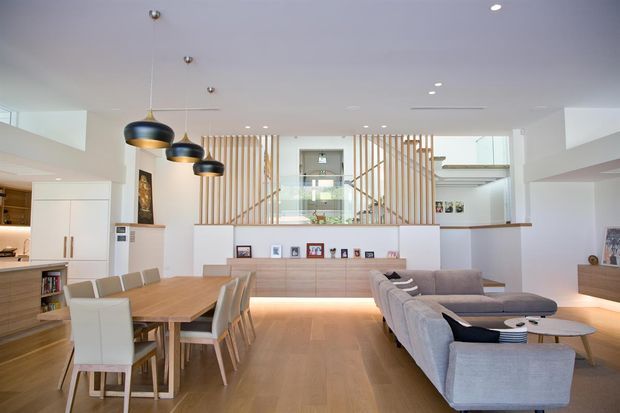 Bright open-plan contemporary interior combining a dining area and living room. The space features pale timber flooring, white walls and cabinetry, a large timber dining table, upholstered dining chairs, a grey sectional sofa, layered pendant lighting, recessed downlights, and a timber slat screen partially separating the upper-level stair or landing area. Built-in shelving and display niches add definition to the room.