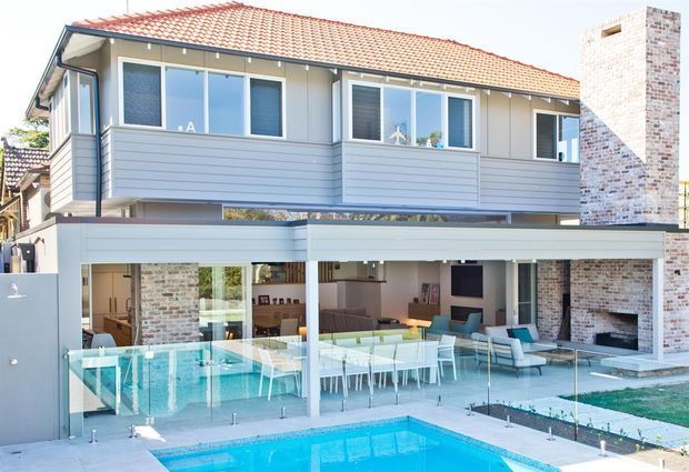 Exterior view of a two-storey contemporary home with a terracotta tiled pitched roof, light horizontal cladding, extensive glazing, and exposed brick chimney and columns. The open lower level connects the living and dining areas to a covered outdoor entertaining space, glass pool fencing, and a swimming pool.