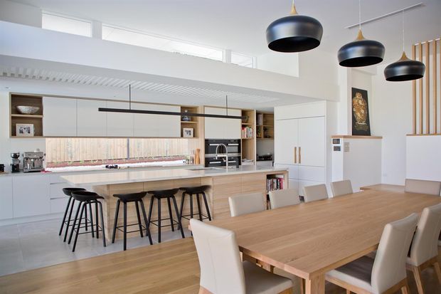 Bright open-plan kitchen and dining area with extensive built-in cabinetry, a long island with bar stools, timber finishes, white surfaces, and a large timber dining table. Sculptural black pendant lights hang above the dining zone, with high clerestory glazing and a partial upper-level void contributing to the airy feel.