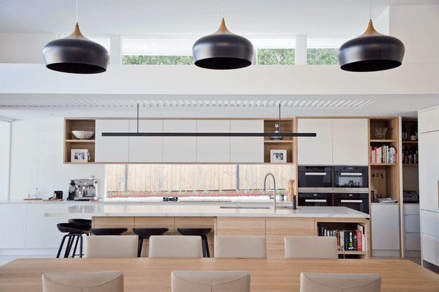 Bright contemporary open-plan kitchen and dining area with extensive white and light timber cabinetry, a central island with seating, integrated appliances, large pendant lights, and a long dining table in the foreground. A timber-toned splashback and open shelving add warmth, while clerestory glazing and a slatted ceiling detail bring in natural light.