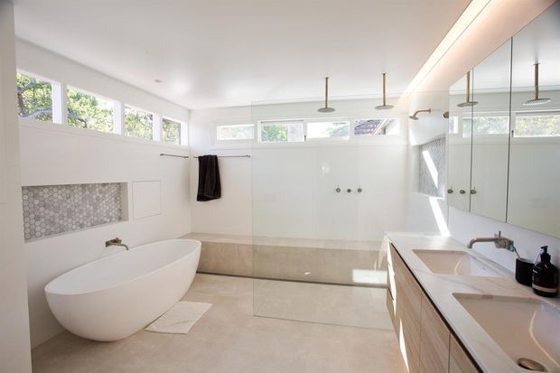 Bright, spacious contemporary bathroom with a freestanding white bathtub, large walk-in glass shower, double vanity with pale timber cabinetry, mirrored wall cabinetry, neutral tiled surfaces, and high clerestory windows. The room has abundant natural light and a clean, minimal finish.