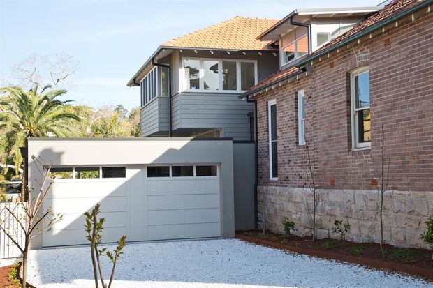 Exterior view of a residential home combining an older brick-and-stone masonry section with a contemporary grey-clad upper extension. The property includes a tiled pitched roof, white double garage, gravel driveway, and mature landscaping. The image highlights the contrasting materials and the integration of new construction with the existing house.