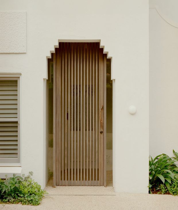 Close-up exterior view of a residential entry facade featuring a tall timber slatted front door set within a stepped geometric arched surround. The facade is finished in warm off-white textured plaster, with a louvred window to the left, simple round wall light, and low tropical planting at the base. The entrance has a refined, warm contemporary character with strong vertical detailing.