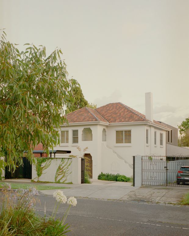 Street-facing view of a suburban two-storey rendered masonry house with a terracotta tiled hipped roof, white-framed louvred windows, exterior chimney, front driveway and metal gates. Mature planting partially obscures the facade, with a neighbouring structure visible at the side.