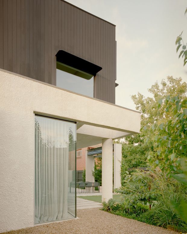 Exterior architectural view of a contemporary two-storey residence, showing a textured pale stucco lower level with a deep covered terrace, slender round column, large glazed opening, and landscaped courtyard. The upper level is clad in vertically ribbed dark timber or composite boards and includes a projecting black-framed window with a green-toned awning. Gravel paving, planting, and glimpses of an outdoor dining area reinforce the courtyard setting.