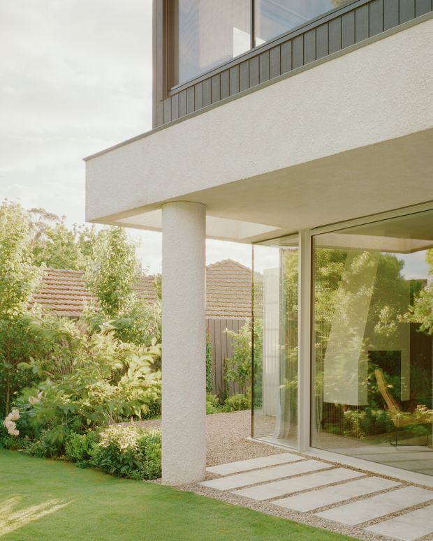 Exterior architectural view of a contemporary residence, showing a projecting upper-level balcony or terrace with a textured white rendered soffit and fascia, supported by a round white column. Full-height glazed doors and windows open onto a landscaped garden with gravel paving and large rectangular stepping slabs. A dark vertical-panel balustrade and timber-framed upper glazing add contrast, while neighbouring tiled roof forms are visible in the background.